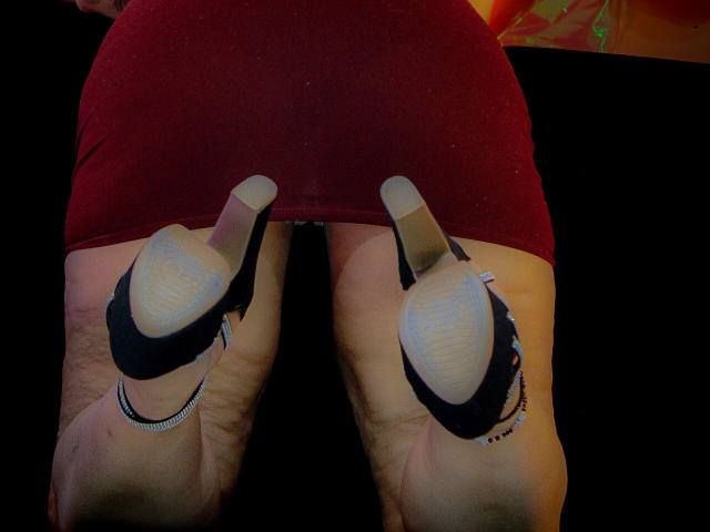 Mariehss's ass in a tight red skirt, wearing black high heels with beige soles, legs slightly apart, showing a bracelet on each ankle, in a dimly lit setting.
