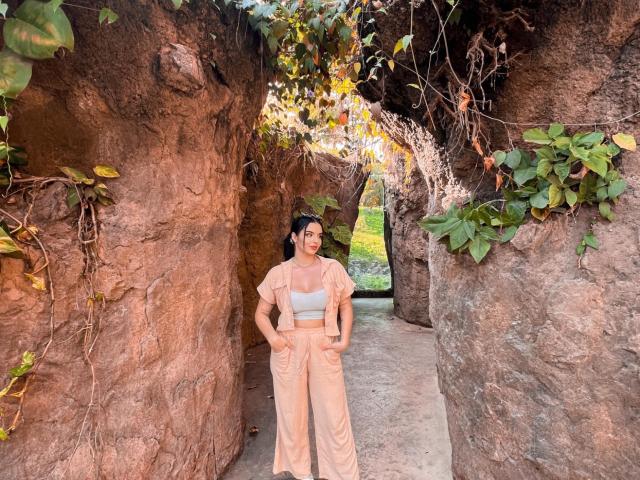 NovaDixon stands in a rocky, vine-covered passageway wearing a beige crop top and matching pants, with hands in pockets, brown hair tied back, looking forward with a calm expression.