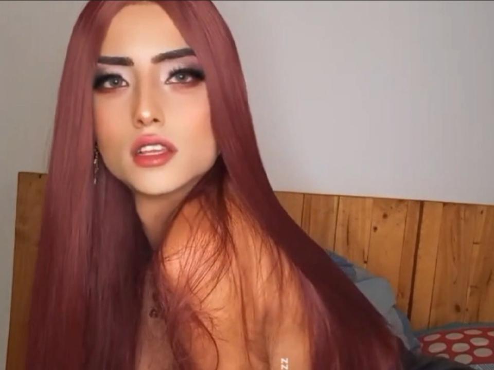 ChristineSapphire with long red hair, heavy makeup, and slightly parted pink lips, looking over her shoulder with a sultry expression, topless, showing her bare shoulders and small nipple.