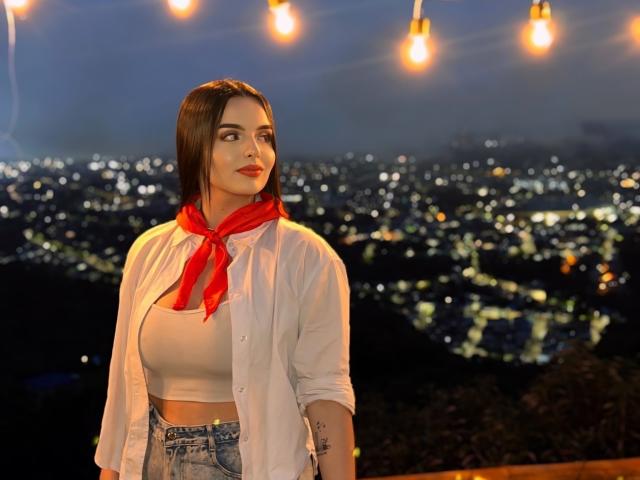 NovaDixon with red scarf, white shirt, and crop top, showing her big boobs and tattoo, standing under string lights, city lights in background, night, dark sky.