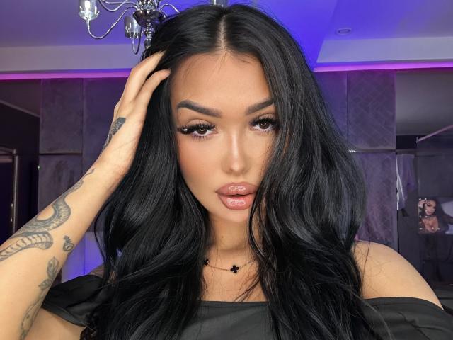 SashaFerrera with long black hair, full lips, and tattoos on her right hand, wearing black off-shoulder top, looking at camera, hand in hair, blue purple lighting.