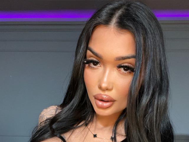 SashaFerrera has long black hair, glossy brown eyes, and full pink lips. She's wearing eyeliner and a black halter top, with a star necklace. Her ass is not visible.
