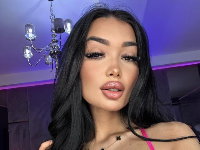 SashaFerrera with glossy pink lips, long black hair, thick eyebrows, and long eyelashes, wearing a pink top, in a room with purple lighting and a chandelier.