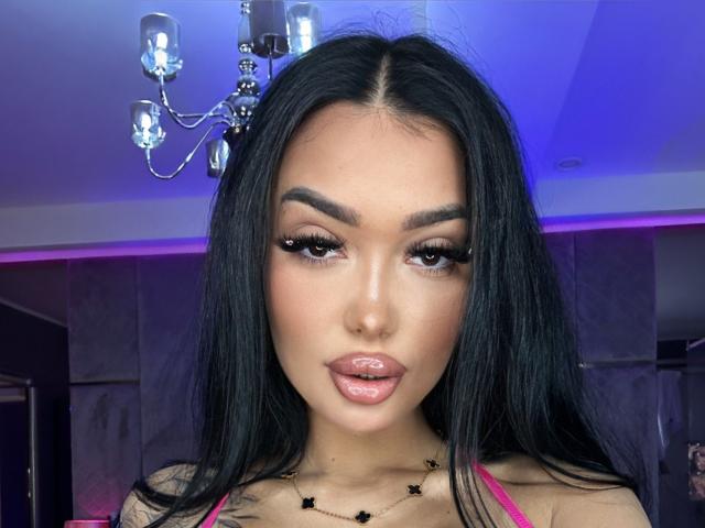 SashaFerrera has long black hair, full eyebrows, thick eyelashes, glossy pink lips, and a necklace with black stars. She's looking at the camera with a seductive expression.