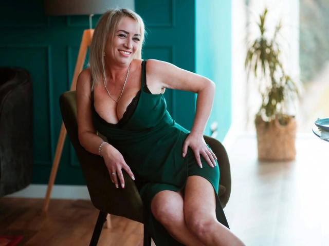 Blonde MilfPamela-hot in green dress, showing her big boobs, smiling, sitting on chair, hands on knees, wooden floor, lamp, and potted plant in background, looking sexy.