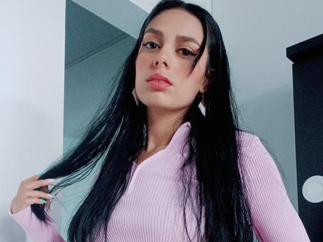 AnaisHilton with long black hair, pink ribbed sweater, pink lipstick, and white nails, holding her hair, looking at the camera with a serious expression.