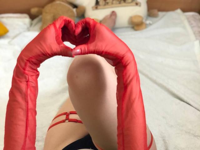 Vailett's pale legs on a bed, wearing red strappy lingerie and red latex gloves making a heart shape with fingers, showing a red-painted nail.