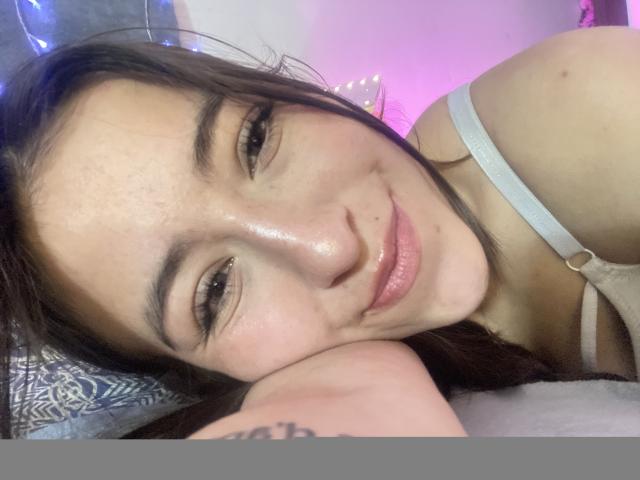 KatiaRogers with shiny skin, brown eyes, and a slight smile, lying down with her head on a pillow, wearing a white bra, tattoo on her arm.