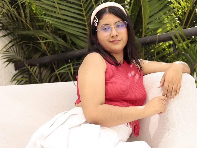 TereCarpert avec une poitrine de taille moyenne, portant un haut rose côtelé, une jupe blanche et un serre-tête rose, avec des lunettes, assise sur un canapé blanc, entourée de plantes vertes..