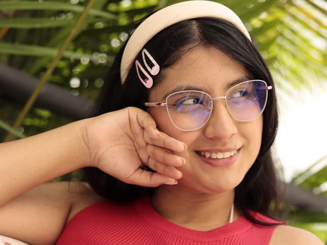TereCarpert has a cute smile, wearing pink cat-eye glasses, a beige headband with pink clips, and a red sleeveless top. Her hand rests on her cheek.