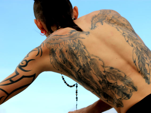 YeremyWalker está de volta, com as costas cobertas por tatuagens detalhadas de penas pretas, com um design tribal no braço direito, usando contas pretas no pescoço, sob um céu azul claro..