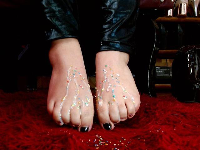 LexyDom's feet with black nail polish, covered in colorful glitter, resting on a red shaggy carpet, wearing black leather pants, showing off her shiny soles.