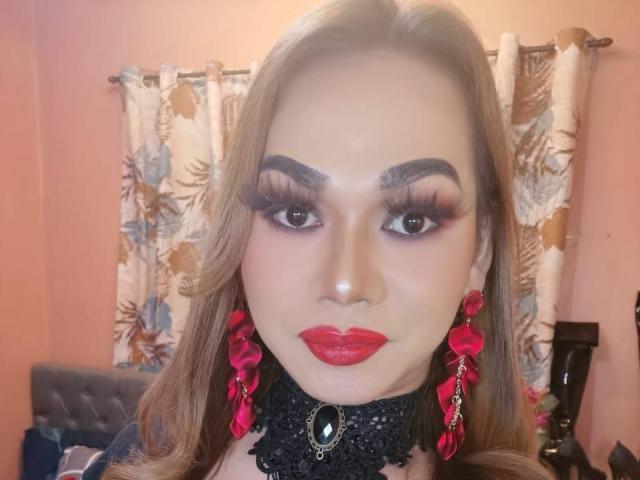 MarianeOhara hat langes, braunes Haar, roten Lippenstift, viel Make-up mit langen Wimpern, eine schwarze Spitzenkette und rote, blumige Ohrringe. Sie blickt mit ernstem Gesichtsausdruck in die Kamera..