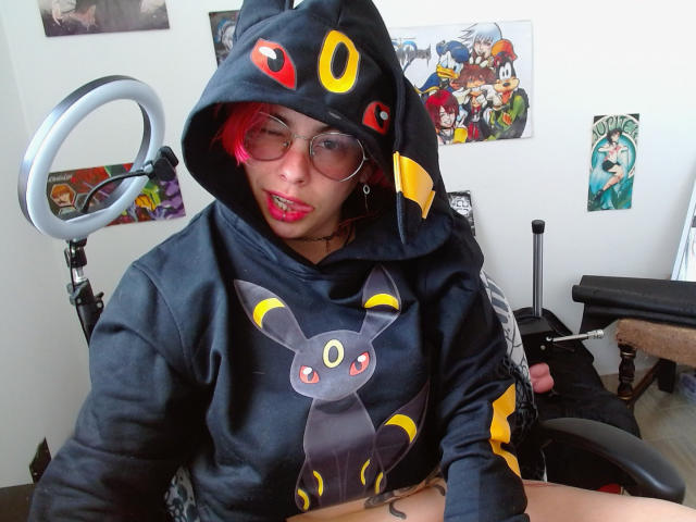 NatashaLau with pink hair, glasses, and a black Umbreon hoodie, mouth open, ring light on left, chair in front, posters on white wall, visible tattoo on right wrist.