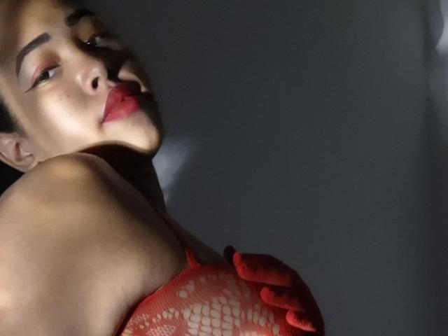 LixAmapola with red fishnet top, covering her tits with red-gloved hands, shadowed face, red lips, looking to the side, dark background, soft light, subtle confident expression.