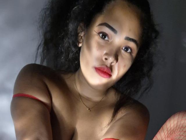 LixAmapola has curly black hair, brown skin, red lipstick, and is topless with a red strap, wearing a gold necklace, looking at the camera with a pouty expression.