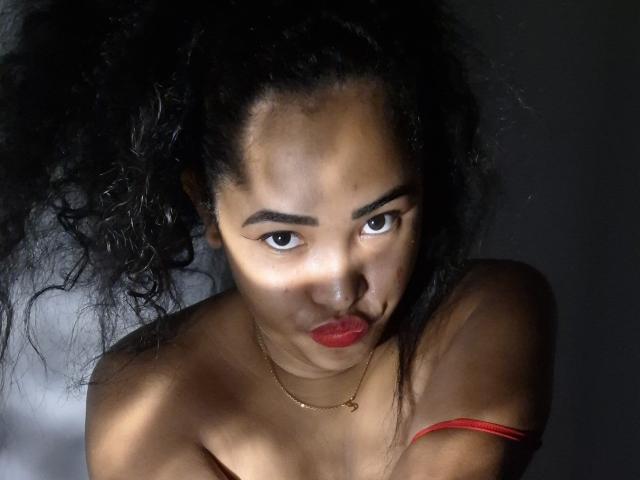LixAmapola has dark curly hair, brown skin, and red lipstick. She's topless with a red strap, looking up with a slight pout. She wears a gold necklace.