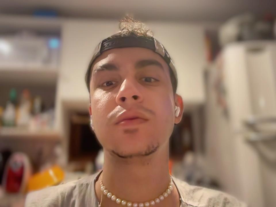 LatinoHottGuy69 mit einer schwarzen Baseballkappe, einem kleinen Bart, einer Perlenkette, weißen Kopfhörern und einem leichten Lächeln, der direkt in die Kamera schaut, in einem unscharfen Innenraum..
