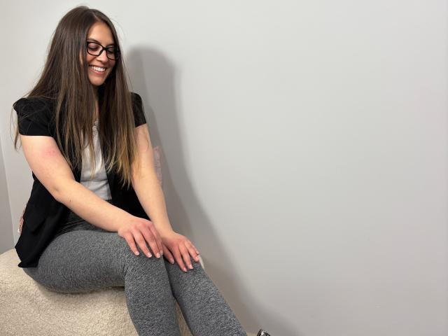 AnnaFlower69, com cabelos castanhos longos, óculos e um sorriso, está sentada em um tapete branco, vestindo uma camisa preta, uma camiseta branca e leggings cinzas, exibindo seus seios e nádegas..