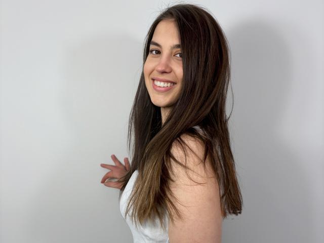 AnnaFlower69 tem cabelos castanhos longos, está sorrindo mostrando dentes brancos, veste uma blusa branca sem mangas, expondo seus seios, e tem uma tatuagem na mão direita, tudo isso em um fundo simples..