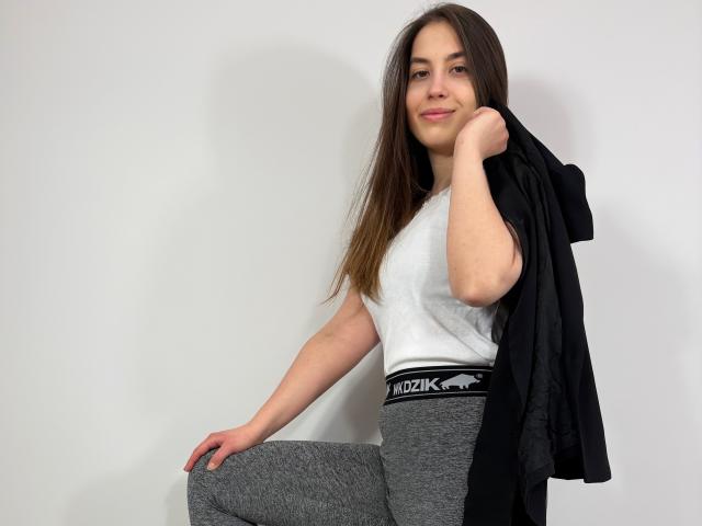 AnnaFlower69, com cabelos castanhos longos, camiseta branca, calças cinzas HooKi, jaqueta preta sobre o ombro, sorrindo, mão no joelho, fundo branco, em pé, relaxada, confiante, fofa..