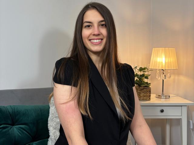 AnnaFlower69, souriante, avec de longs cheveux bruns, portant une robe noire, assise sur un canapé vert, une petite table blanche avec une lampe et une plante, elle a l'air mignonne et ses seins sont visibles..