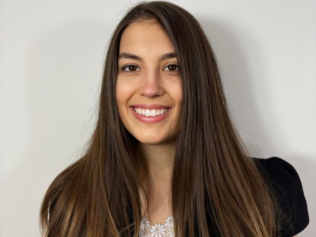 AnnaFlower69 tem cabelos castanhos longos, olhos castanhos e um sorriso que mostra dentes brancos. Ela está vestindo uma blusa preta com um padrão de renda branca por baixo..