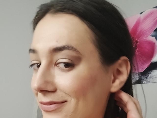 JuliaHuntis has a smug smile, dark brown eyes, and a small mole on her left cheek. She's holding her hair back with a hand. Her tits are not visible.