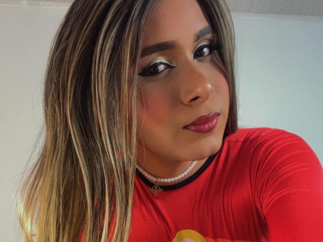 NattyTrans has long blonde and brown hair, wearing a red shirt, pearl necklace, and red lipstick, with black eyeliner and subtle makeup, smiling at the camera.