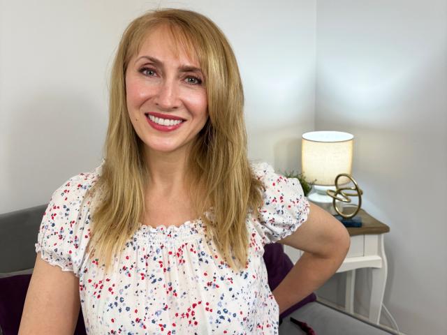 MargaretPretty69 with blonde hair, smiling, wearing a white floral top, hands on hips, sitting next to a lit table lamp with a brass base.