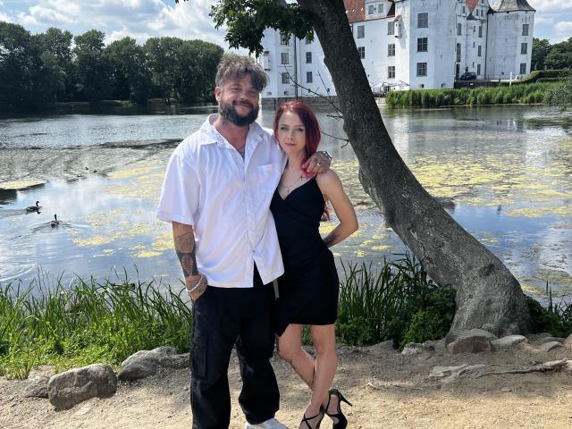 WeGoHard, con cabello rojo y vestido negro, de pie junto a WeGoHard, con barba gris y camisa blanca, ambos cerca de un estanque con una casa blanca al fondo..