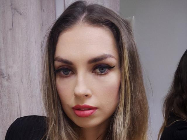 SiennaPearly has blue eyes, thick black eyebrows, long eyelashes, pink lipstick, and blonde hair with darker roots, wearing a black top. Her face is centered in the photo.