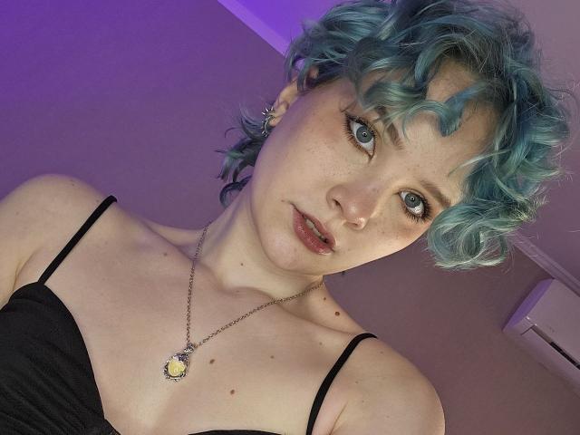 ShellyLove69 has blue curly hair, fair skin, freckles, and blue eyes. She wears a black spaghetti strap top and a necklace with a yellow pendant. Her tits are small.