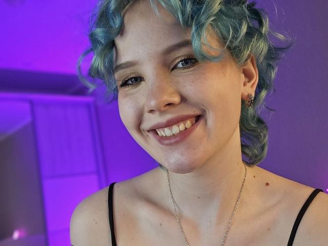 ShellyLove69 with blue curly hair, smiling, showing teeth, wearing a black spaghetti strap top, small earrings, and a silver necklace, with visible freckles and a cute nose.