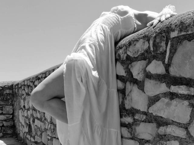 CocoMelle is lying on a stone wall, her dress pulled up, exposing her bare ass. She's wearing a white dress, and the image is black and white.