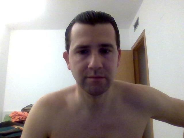 JorgeLih, shirtless, with short dark hair, slight beard, and a neutral expression, standing in a room with a wooden door and some clothes on a bed.