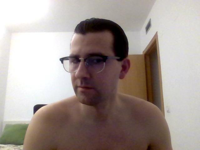 Shirtless JorgeLih with glasses, short dark hair, slightly parted lips, looking down, bare shoulders, light skin, wooden door in background, simple white room, slightly blurry image.