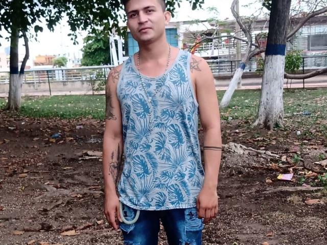 Cirio, wearing a blue tropical-patterned tank top and jeans, stands in a park with tattoos on both arms, looking slightly bored. He has a chain necklace and short hair.