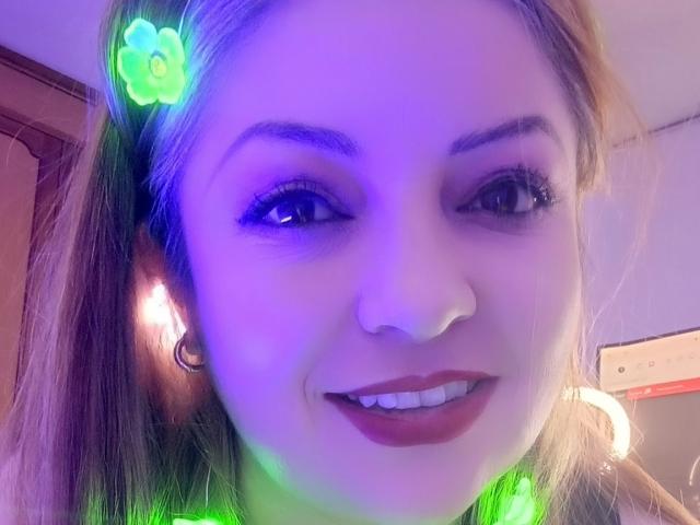 NatalyHami with a green clover hair clip, smiling, wearing hoop earrings, purple and green light, brown hair, and red lipstick, in a selfie.
