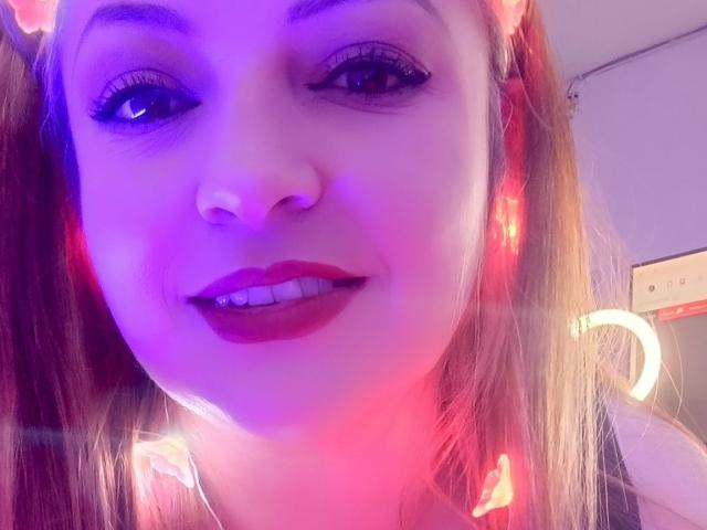 NatalyHami has purple-tinged skin, red lipstick, and long brown hair, smiling with eyeliner, and a nose ring, under pink and blue lights, showing her white teeth.