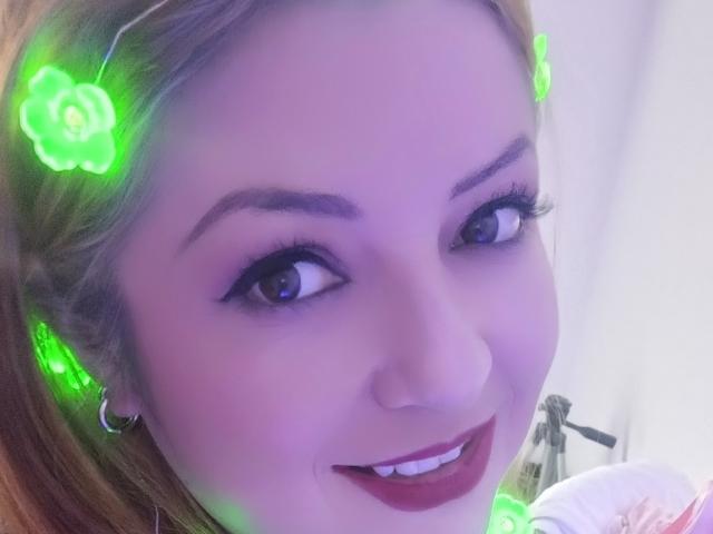 NatalyHami has glowing green flower headbands, purple-tinted skin, and a cute smile with dark lipstick. Her eyes are blue and she's wearing hoop earrings.