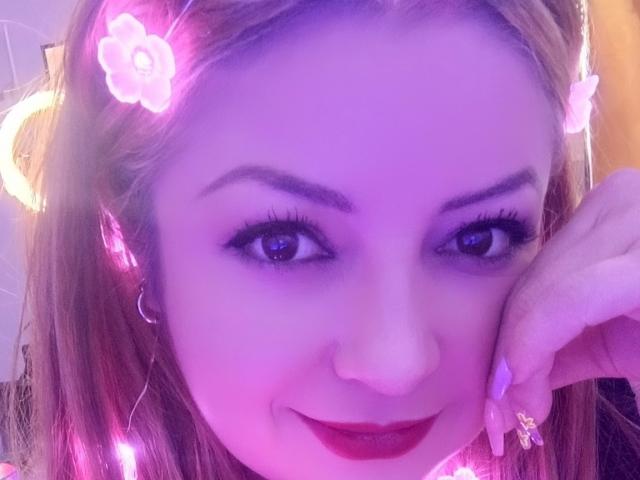 NatalyHami has purple-lit face, red lipstick, long eyelashes, brown hair with pink flower clips, and a cute smile, resting her chin on her hand.