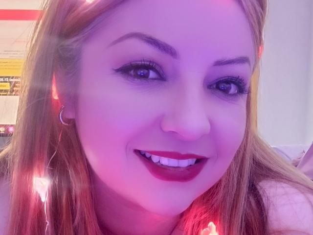 NatalyHami has purple-lit face, long blonde hair, black eyeliner, red lipstick, smiling, showing white teeth, wearing a hoop earring, with a string of red lights in her hair.