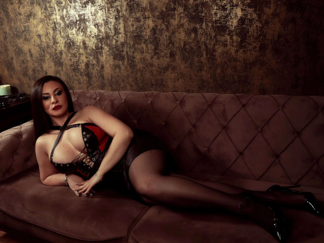 AmberCrost with big tits, lying on a brown couch, wearing black lace lingerie, red accent, black stockings, and black high heels, looking sexy, with dark, wavy hair.