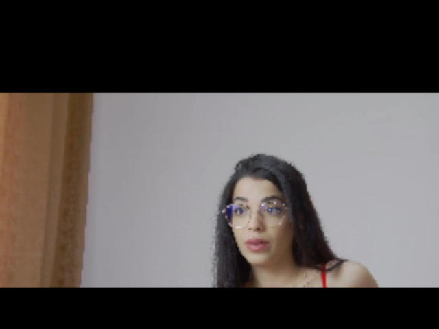 LisaMoon has long black hair, wears glasses, and a red top, with a serious expression, showing her tits slightly, against a plain white background.