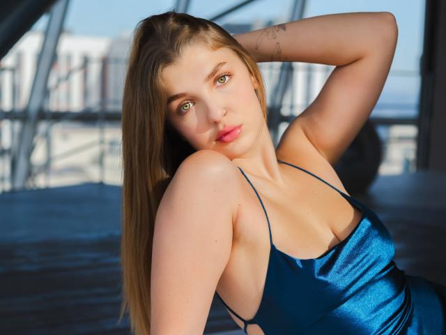 AngelAmalia with green eyes, wearing a blue satin dress, has her arm behind her head, showing her tits and a tattoo on her arm, in a sunlit room.