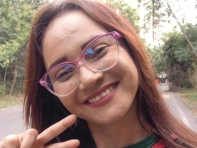 Sofia69s with pink glasses, brown hair, and a smile, making a "peace" sign with her right hand. She's got cute tits and a small nose. Outside on a road.