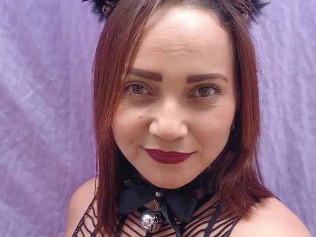 Sofia69s has reddish-brown hair, cat ears, dark lipstick, and a black mesh top with a bell and bow. She's smiling with brown eyes and a small beauty mark.