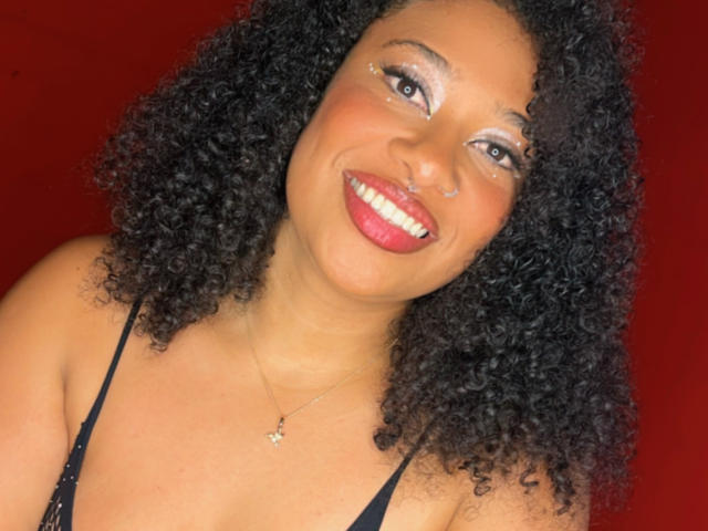 Shanthaly has curly black hair, a nose ring, and is smiling with red lipstick. She's wearing a black top and a small necklace against a red background.