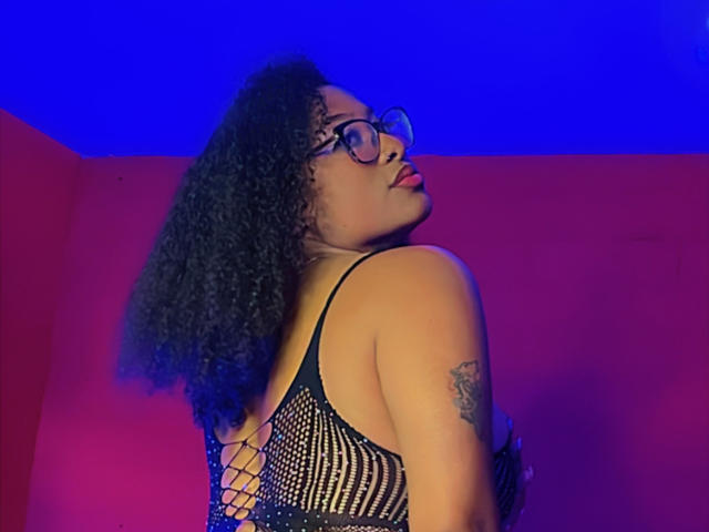 Shanthaly with curly black hair, glasses, and red lipstick, wearing a black mesh dress, showing a tattoo on her right arm, looking back with a sultry expression.